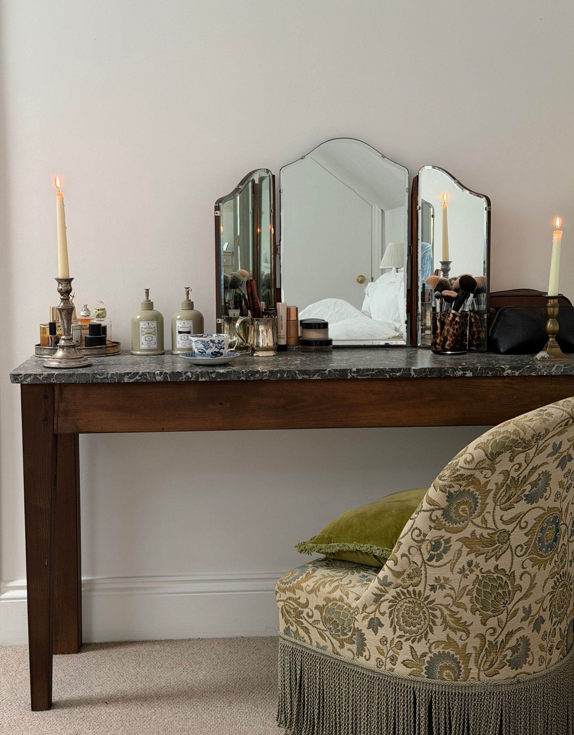 Image of a wooden vanity table with a dark green marble top. The vanity has lots of items on it and a tri-fold mirror. There is a green patterned chair with fringe trim in front.