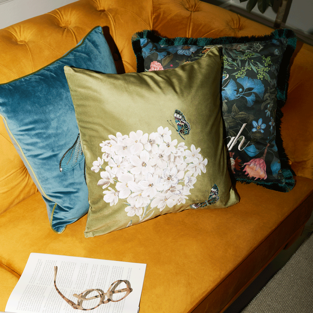 a close up of an orange sofa with blue, green and dark floral patten cushions as well as reading glasses resting on paper on the sofa
