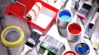 Paintbrushes, paint tins, roller and red roller tray and masking tape arranged in a geometric pattern on a wooden surface