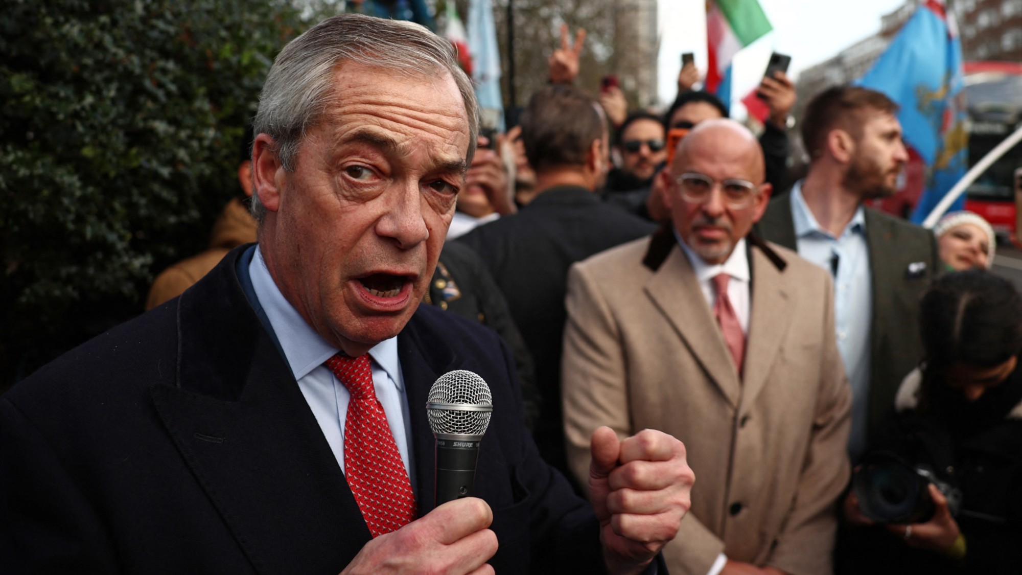 Nigel Farage talks into a microphone, with Nadhim Zahawi behind him