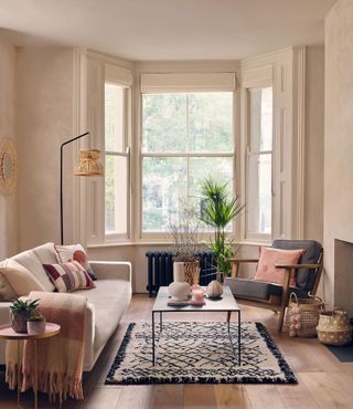Neutral living room with boho, rattan accents, berber high pile rug, black coffee table, cream couch with wool throw, small rose gold side table and gray armchair with small pink cushion