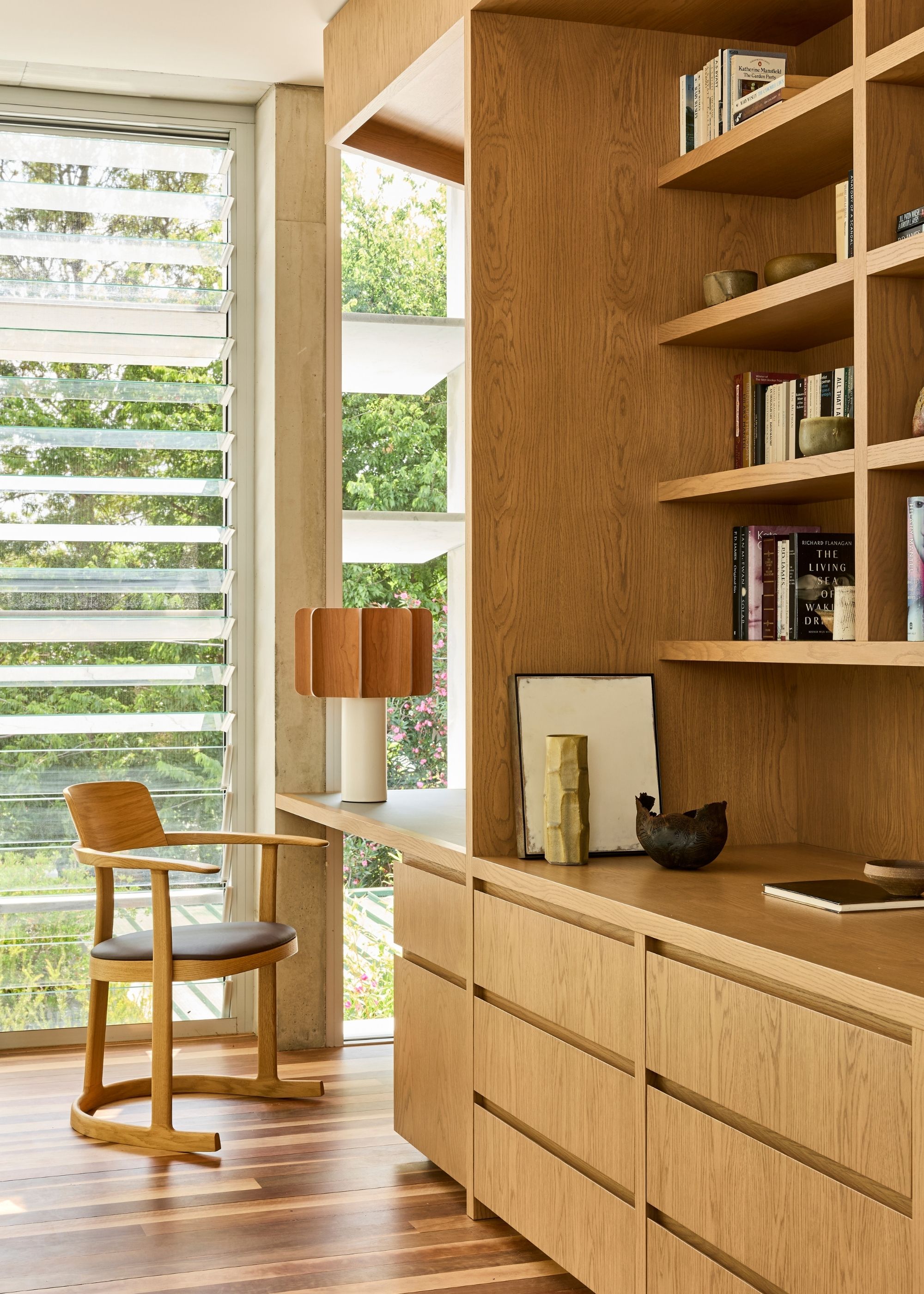 A book display by a small study with a wood chair, a two-toned lamp, and large windows with blinds