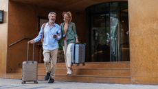 Mature couple joyfully exits a hotel, rolling their suitcases. They appear cheerful and ready for travel adventures