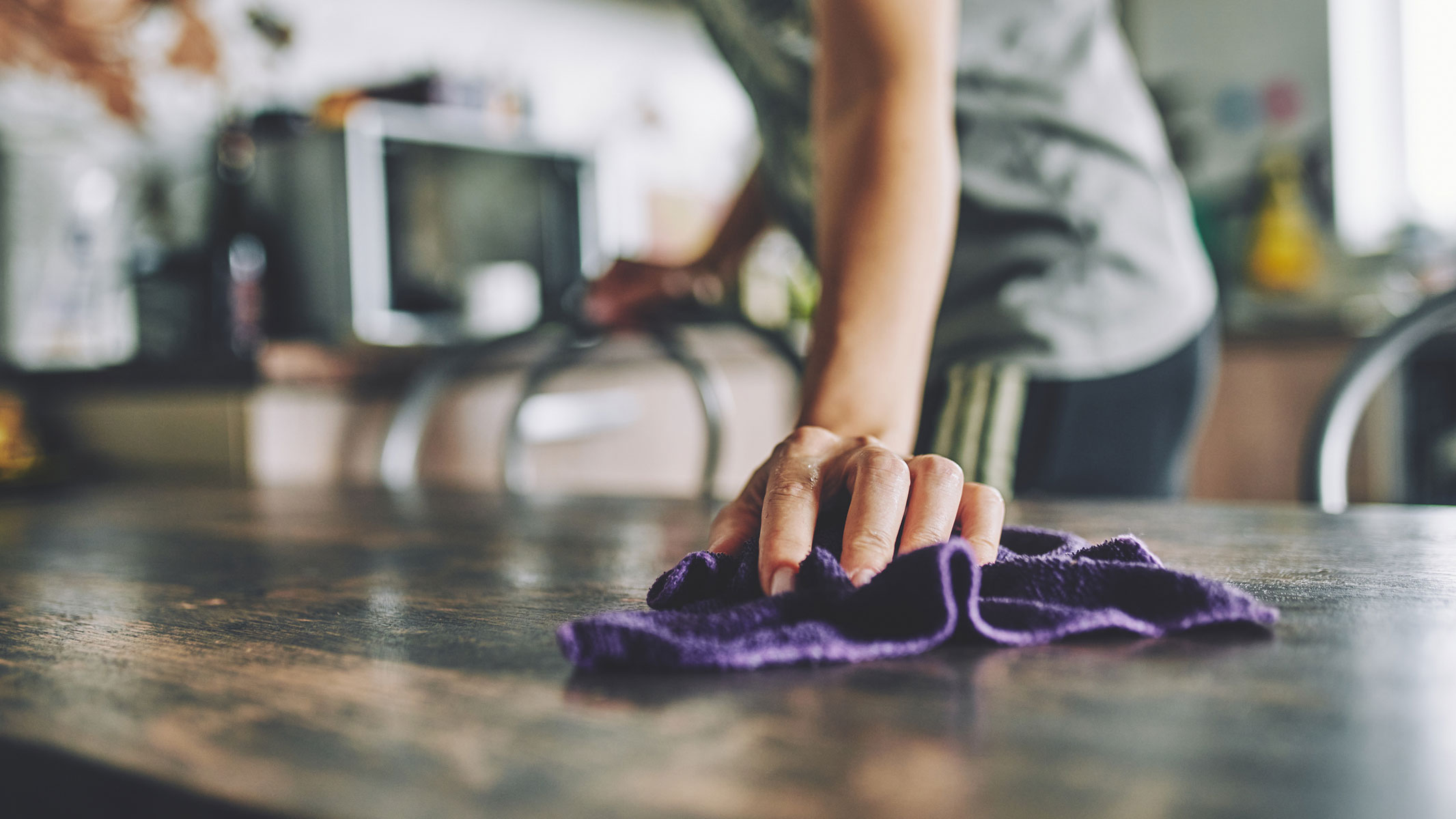 A close-up picture of a woman wiping the kitchen table with a cloth