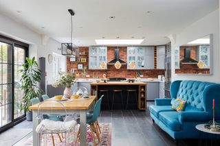 Open-plan kitchen diner with grey Shaker-style kitchen and wooden island, brick slip wall, wooden dining table and chairs on a pink patterned rug, and a large blue velvet sofa