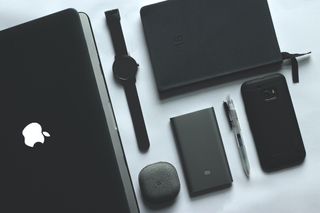 An Apple laptop, smart watch, Samsung phone, pen and notebook, all in black, laid out neatly across a white table