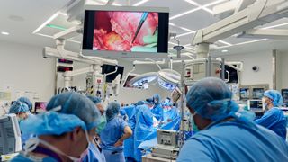 photo of surgeons in blue scrubs, hair nets and masks gathered near an operating table as a monitor shows a kidney being placed into the patient's body