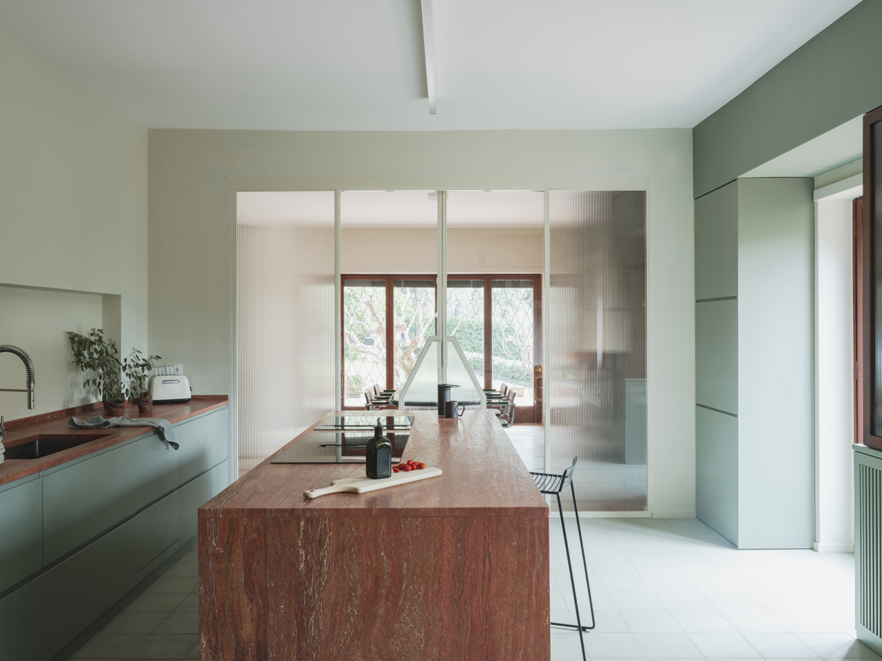 a pale green kitchen with a red travertine island with glass doors dividing it from the dining room