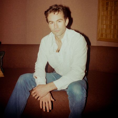 Chace Crawford sits on a brown couch wearing a white button-down shirt and blue jeans. 