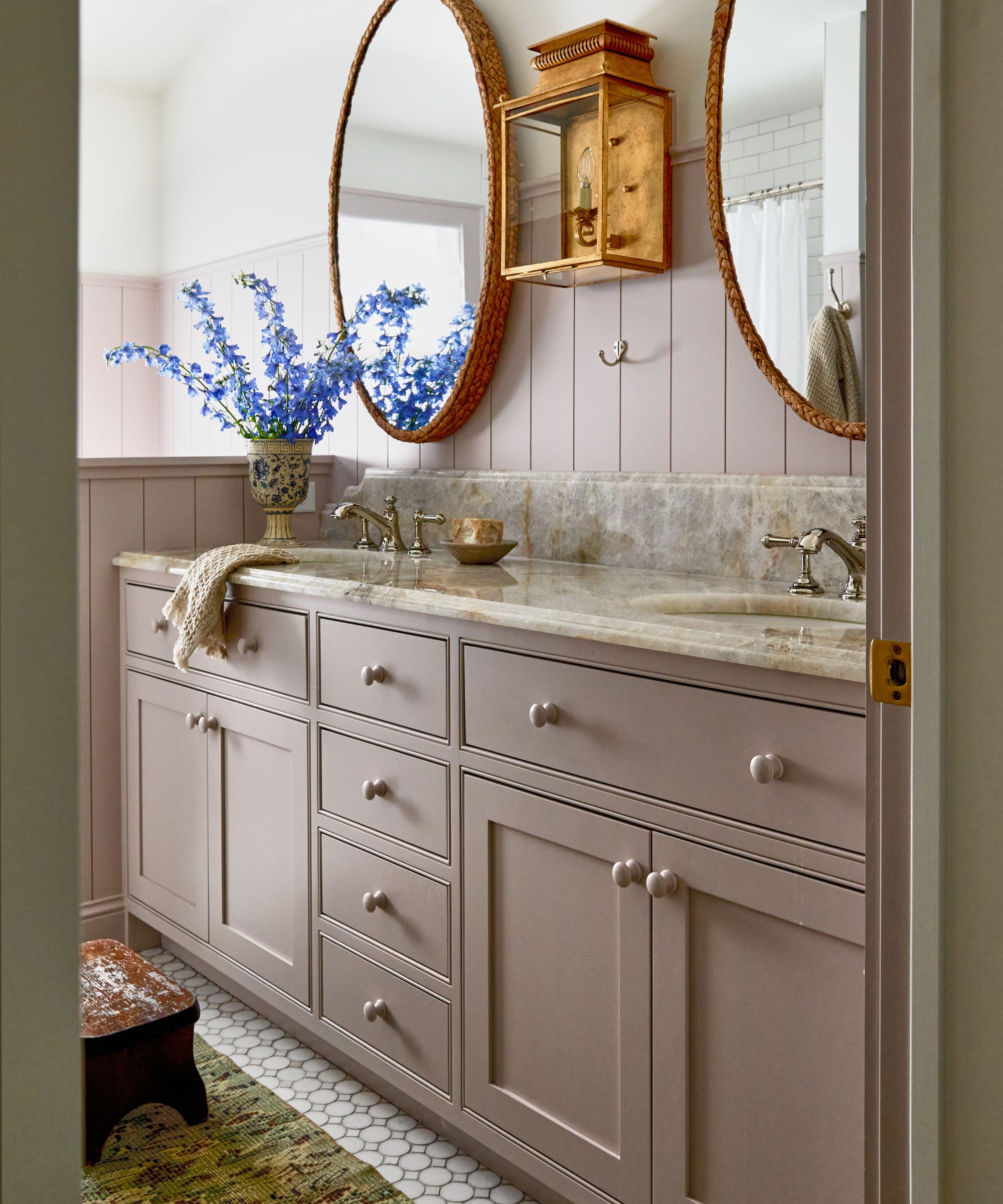 A bathroom with lilac panelled walls and a lilac vanity with double sinks, two oval wall mirrors, a marble countertop, and silver hardware.