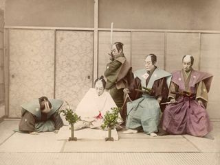 A dramatized photo shows five samurai in a room with tatami mats. Two men are kneeling and three men look at them. One man, who is standing, has a sword.