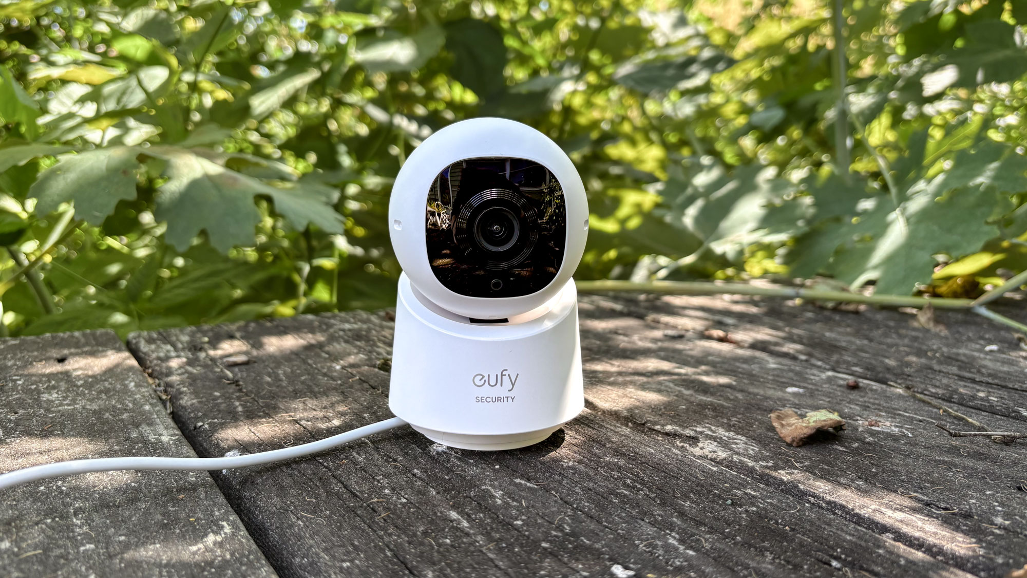 A Eufy home security camera outside on a deck in front of green bushes.
