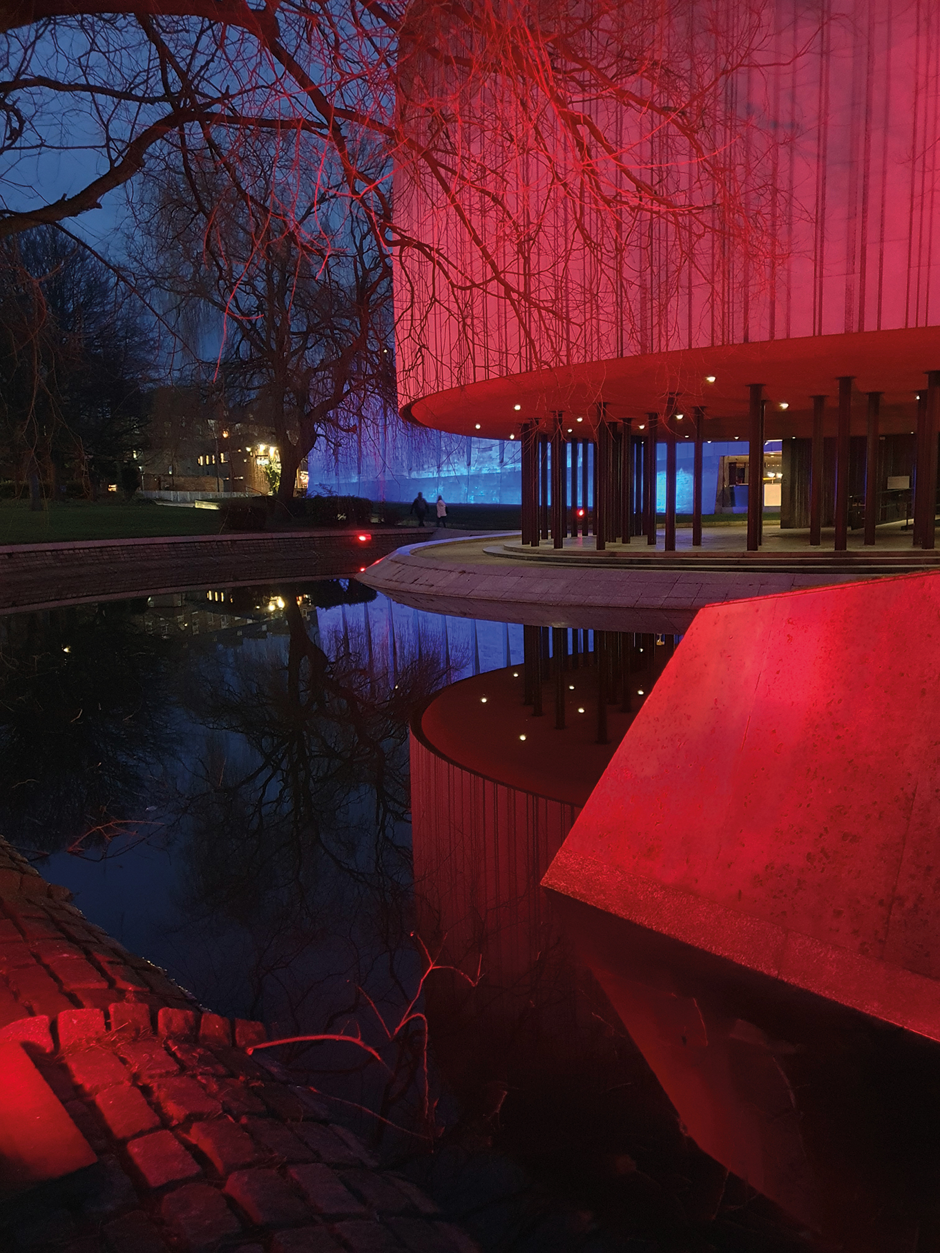 A modern building illuminated in red light reflects in a still pond, framed by trees, under a darkening evening sky