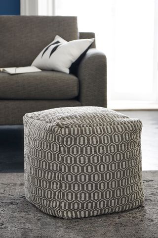 A grey cube shaped bean bag on a living room rug in front of a grey sofa.