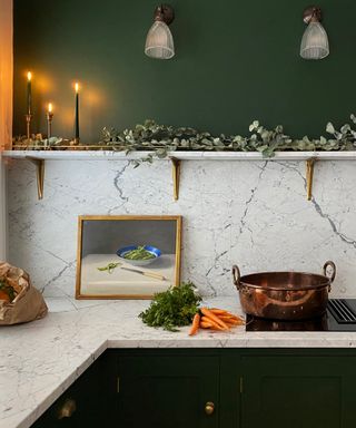 winter decor ideas, green and marble kitchen with small shelf full of eucalyptus, three green candles lit in brass candlesticks, wall lights, large copper pan, carrots, artwork