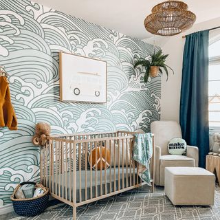 Wooden cot, wavey patterned wallpaper, grey armchair and footstool, navy curtains and splashes of green and yellow