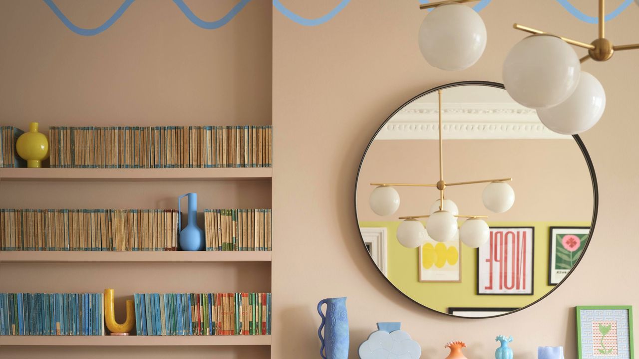 A pale pink living room wall with bookshelves filled with books and a gold circular mirror hanging on it, plus colorful vases below the pillow and a sputnik chandelier in front of it