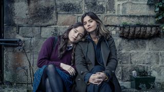 Ruby Stokes in a school uniform as Hannah sits on a bench against a stone wall with Jenna Coleman in a dark coat as Ember in The Jetty