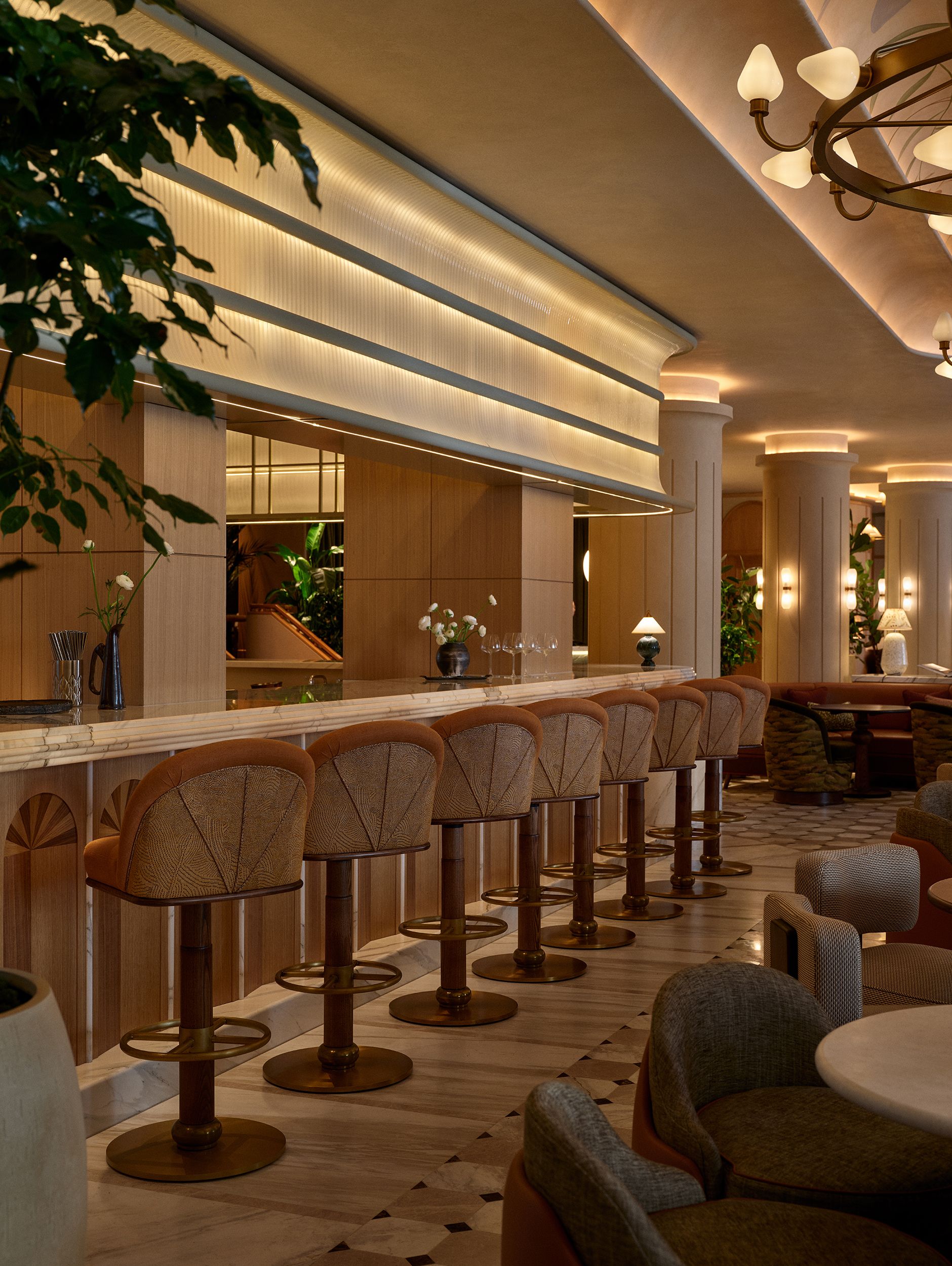 A beautifully decorated hotel bar with a statement mouth-blown glass frieze, upholstered stools in wood and brass, golden lighting, and marble detailing.