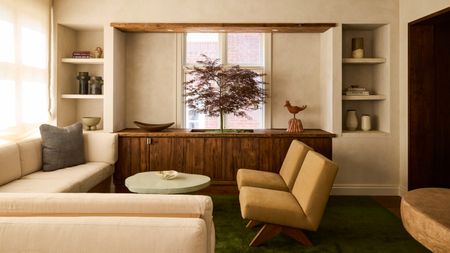 Living room with cream plaster walls, cream corner sofa, ochre armchairs, rough edged mint coffee table, wood sideboard and indoor tree planted inside
