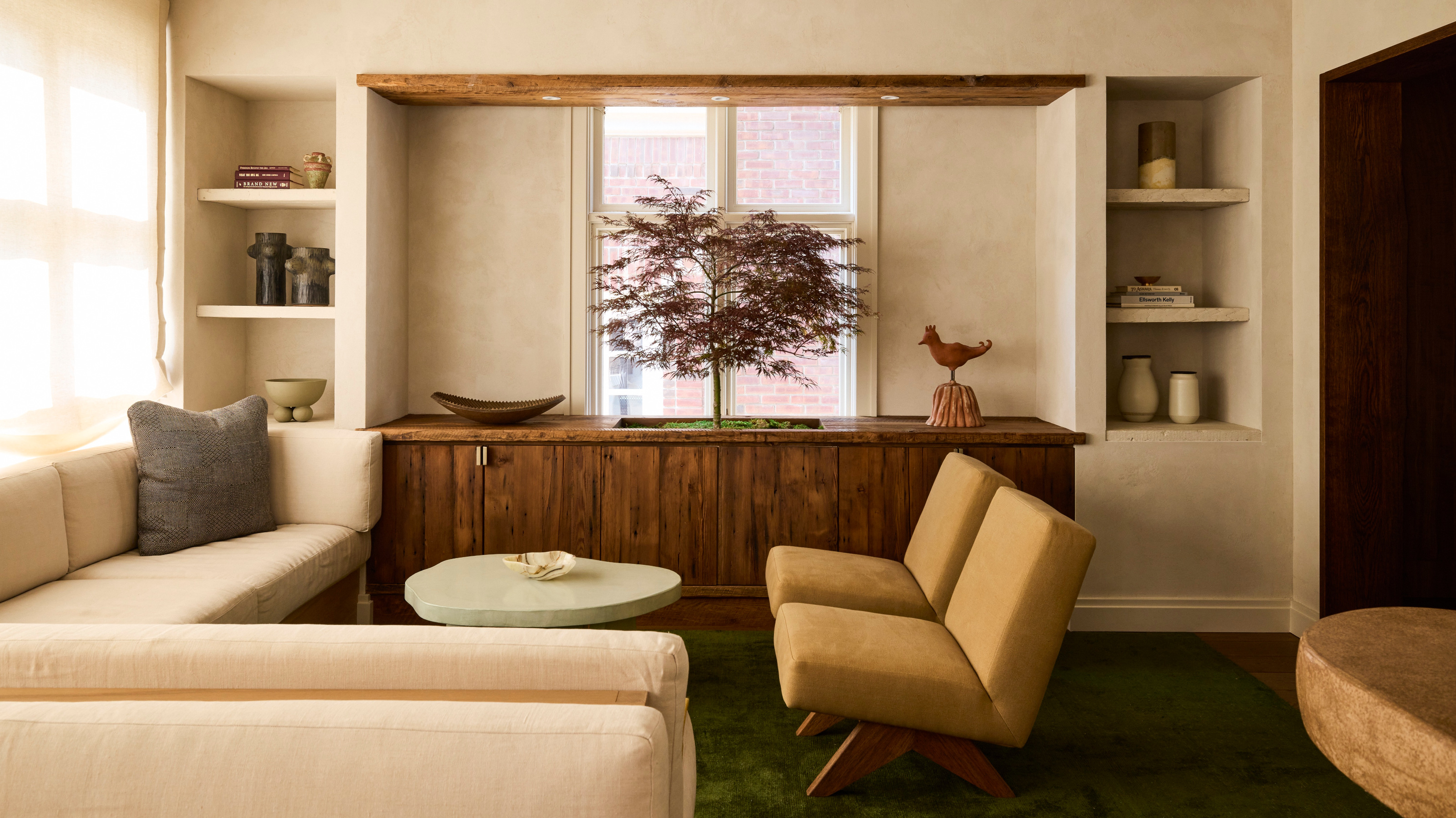 Living room with cream plaster walls, cream corner sofa, ochre armchairs, rough edged mint coffee table, wood sideboard and indoor tree planted inside