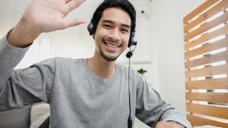 A man wearing a microphone headset and happily waving on a video call