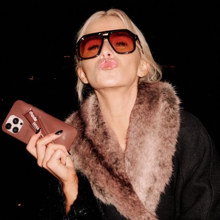 A woman puckers her lips and holds a tube of Rhode lipstick in brown with matching phone case.