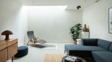 minimalist living room with skylight, plastered walls and ceiling, lounge chair on rug under skylight, timber console with mushroom lamp, blue ottoman on the floor, jute rug under blue L-shaped sofa with black round coffee table, and in the back corner is a plinth with a sculpture on top and potted plant