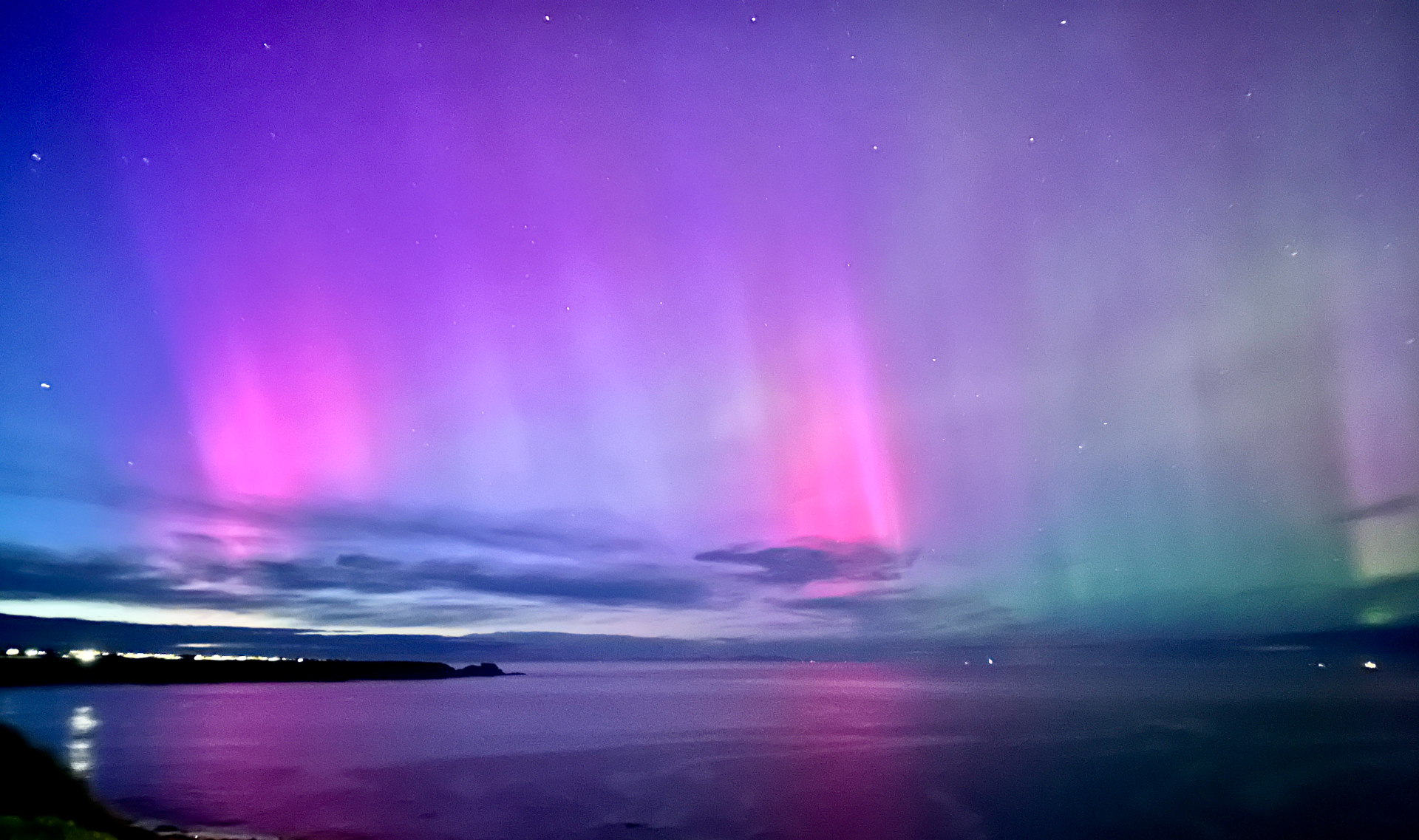 As auroras boreais aparecem em cores vibrantes de rosa e verde como cortinas de luz sobre um grande corpo de água.