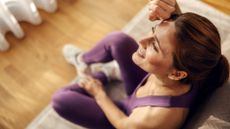 A woman sits on the floor in workout gear, wiping sweat from her head with the back of her hand.