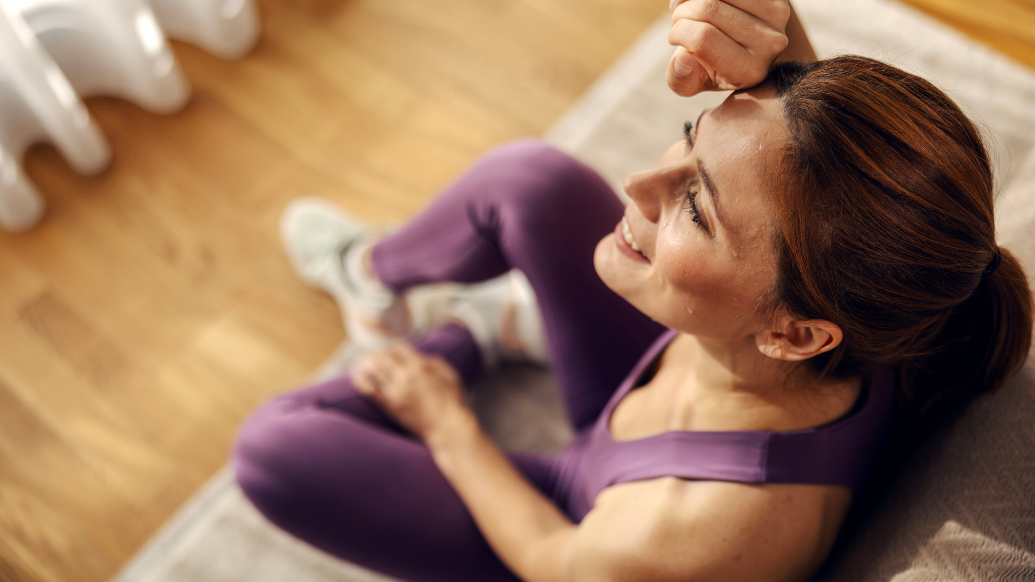 A woman sits on the floor in workout gear, wiping sweat from her head with the back of her hand.