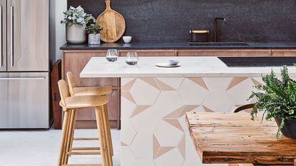 A modern kitchen with American-style refrigerator, island with encaustic tile detail, wooden bar stools and dining table, and black splashback