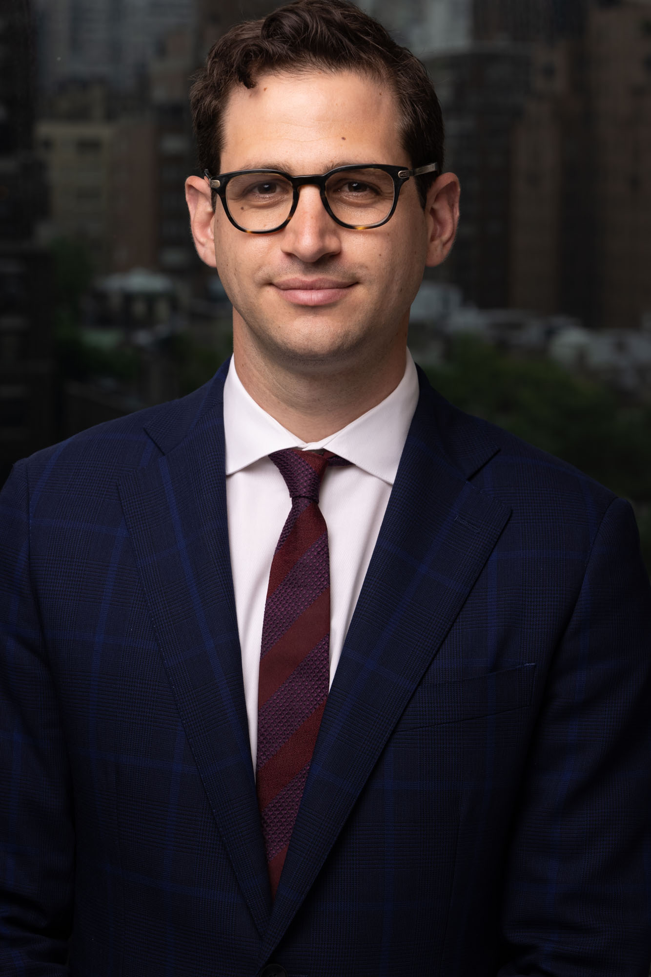 a white man with brown hair in glasses wearing a navy suit and burgundy tie