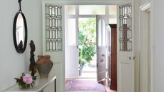 White home entrance with open doorway, patterned red rug, glass paned doors, circular mirror, pink flowers and a terracotta vase