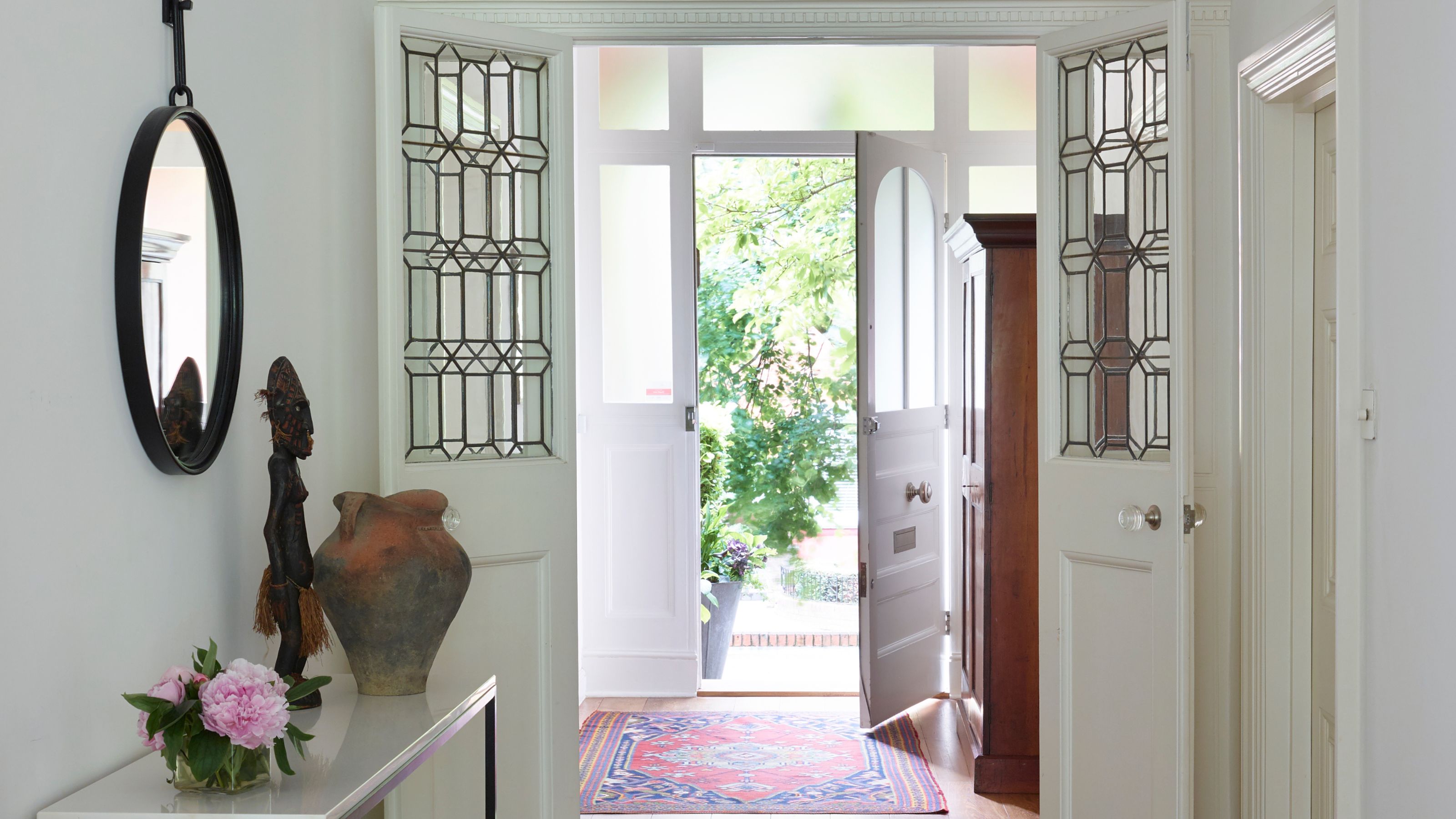 White home entrance with open doorway, patterned red rug, glass paned doors, circular mirror, pink flowers and a terracotta vase