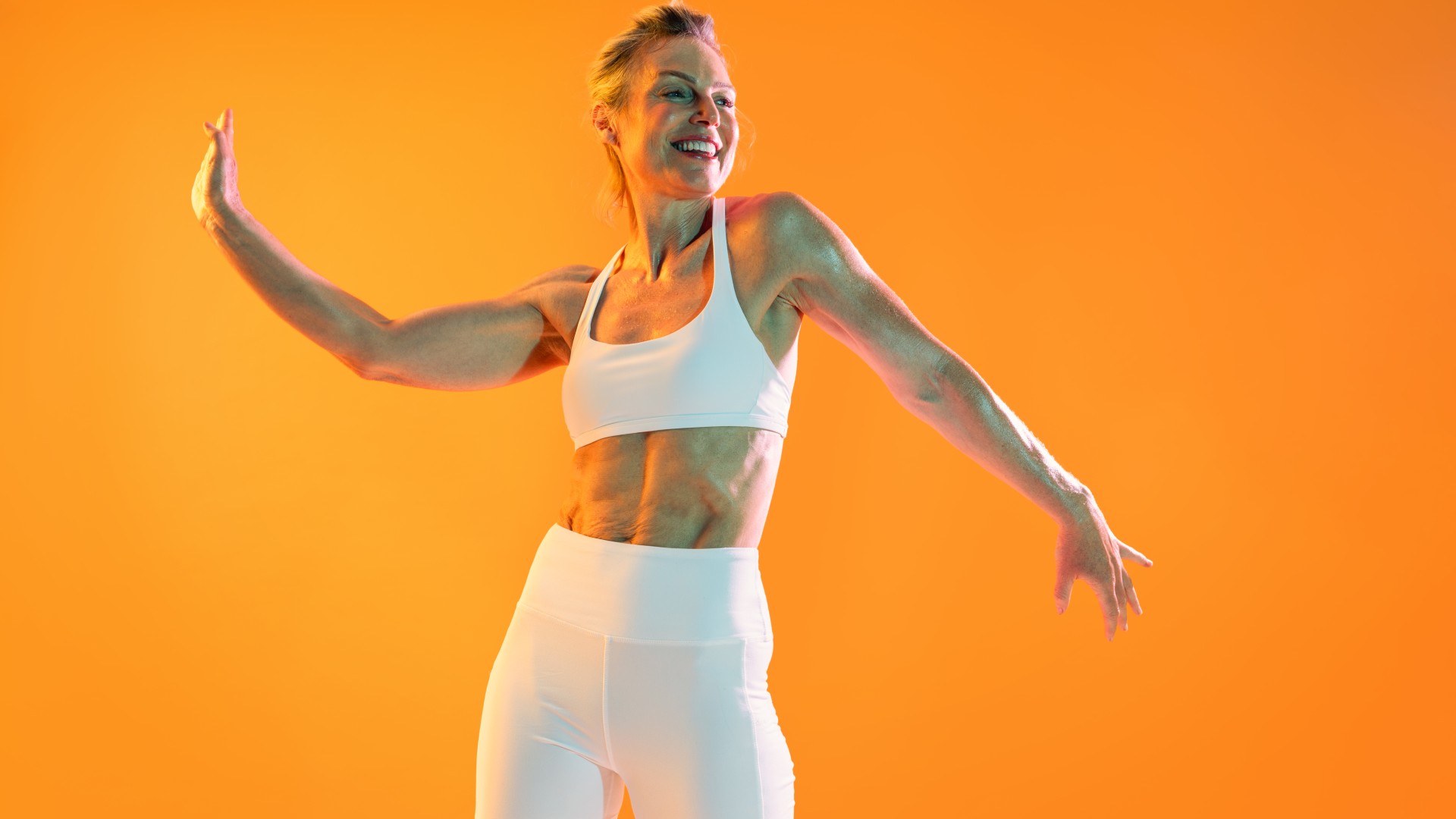 Mature woman in white activewear with abs smiling for camera orange background