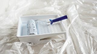 A paint tray and roller with white paint on a floor covered with plastic sheets