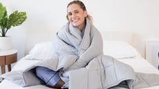 The best weighted blankets in 2022: A woman with blonde hair sits on her bed wrapped in a Luna Adult Weighted Blanket