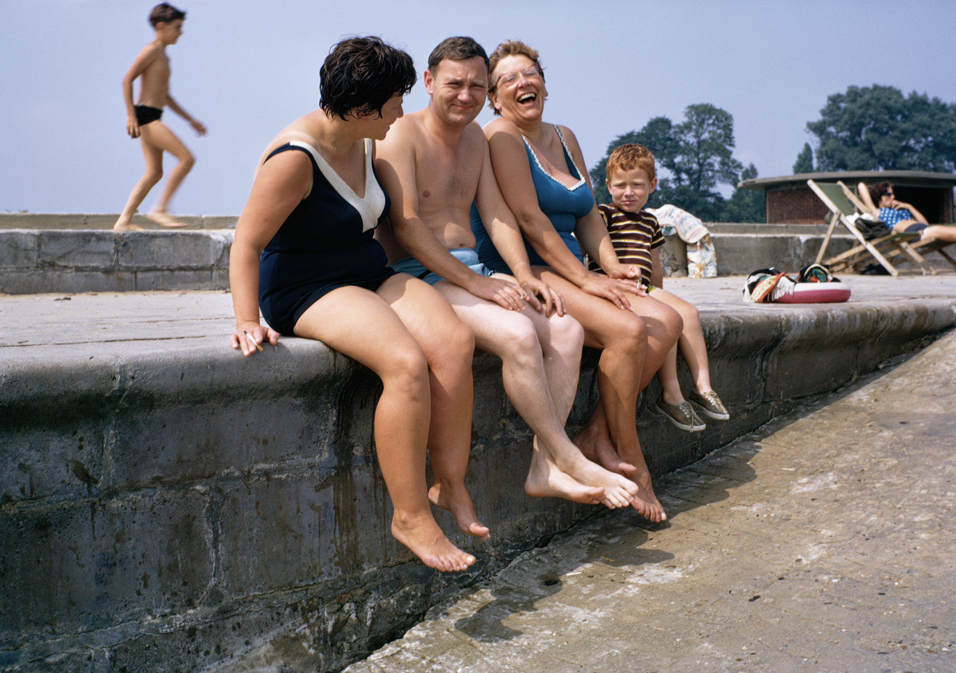 A candid, colorful 1960s photograph capturing a group of adults and a young boy laughing together while sitting with their legs dangling over a concrete sea wall at a public bathing area.