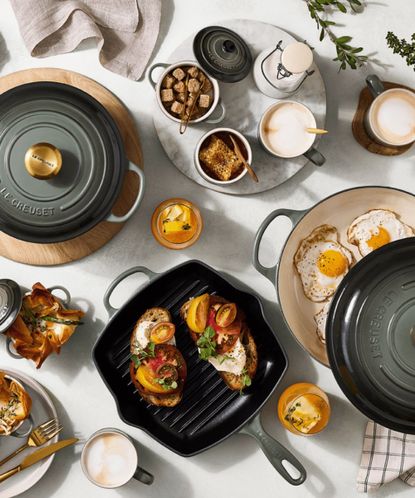 A bird's eye view of breakfast cooking in Le Creuset ovens and griddle pans in Thyme on a white kitchen counter, surrounded by plates and cups. 