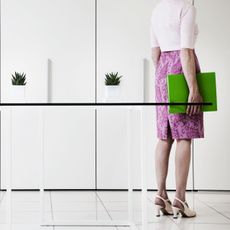 Woman wearing pink skirt, holding green folder, standing in queue in white office by small pot plants