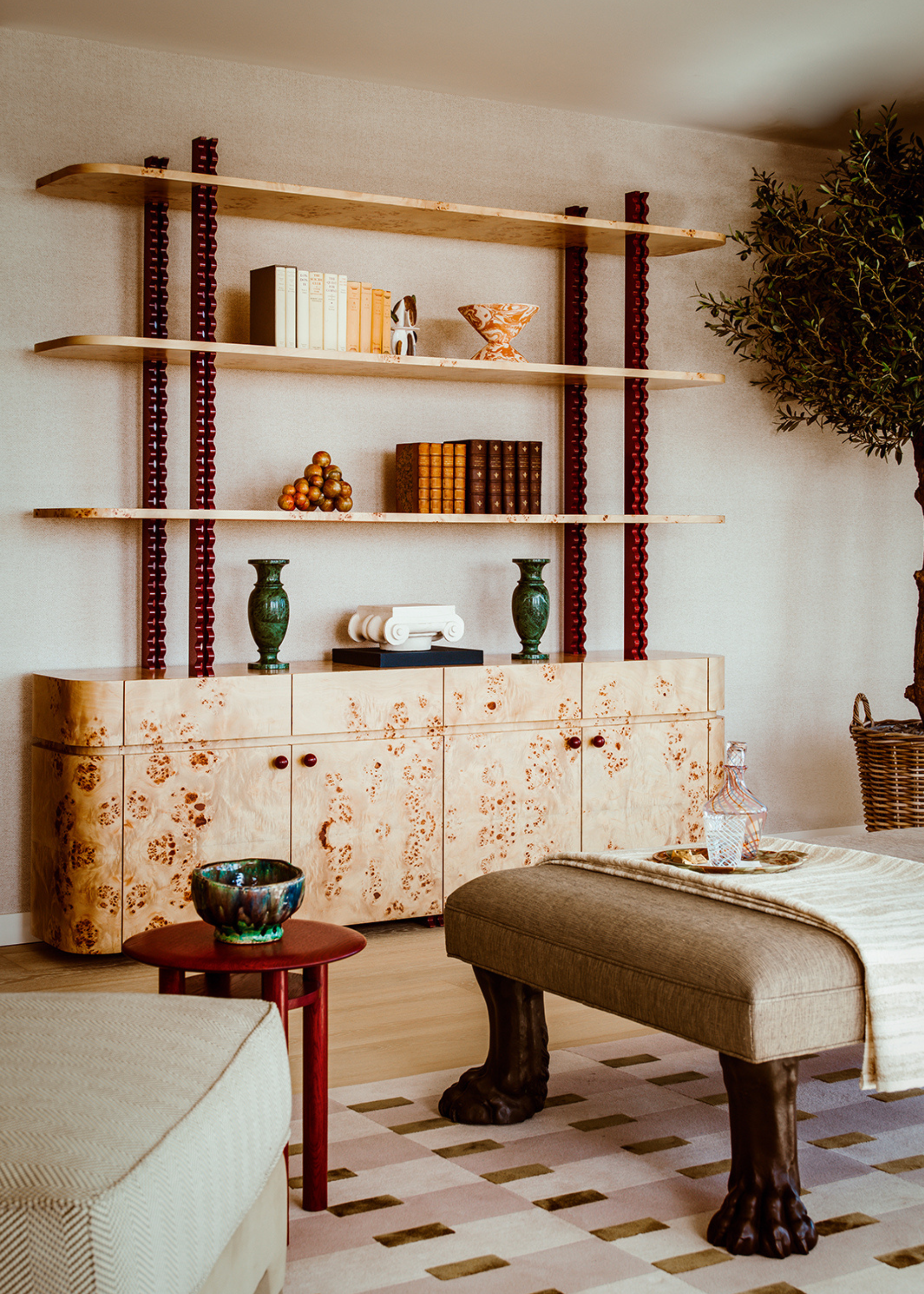 A contemporary living room with neutral walls and a burl wood bookcase of decorative objets with bobbin details on the stands holding up the shelves in a deep red, a large indoor tree on the side and an armchair, ottoman and side table in front on top of a patterned rug