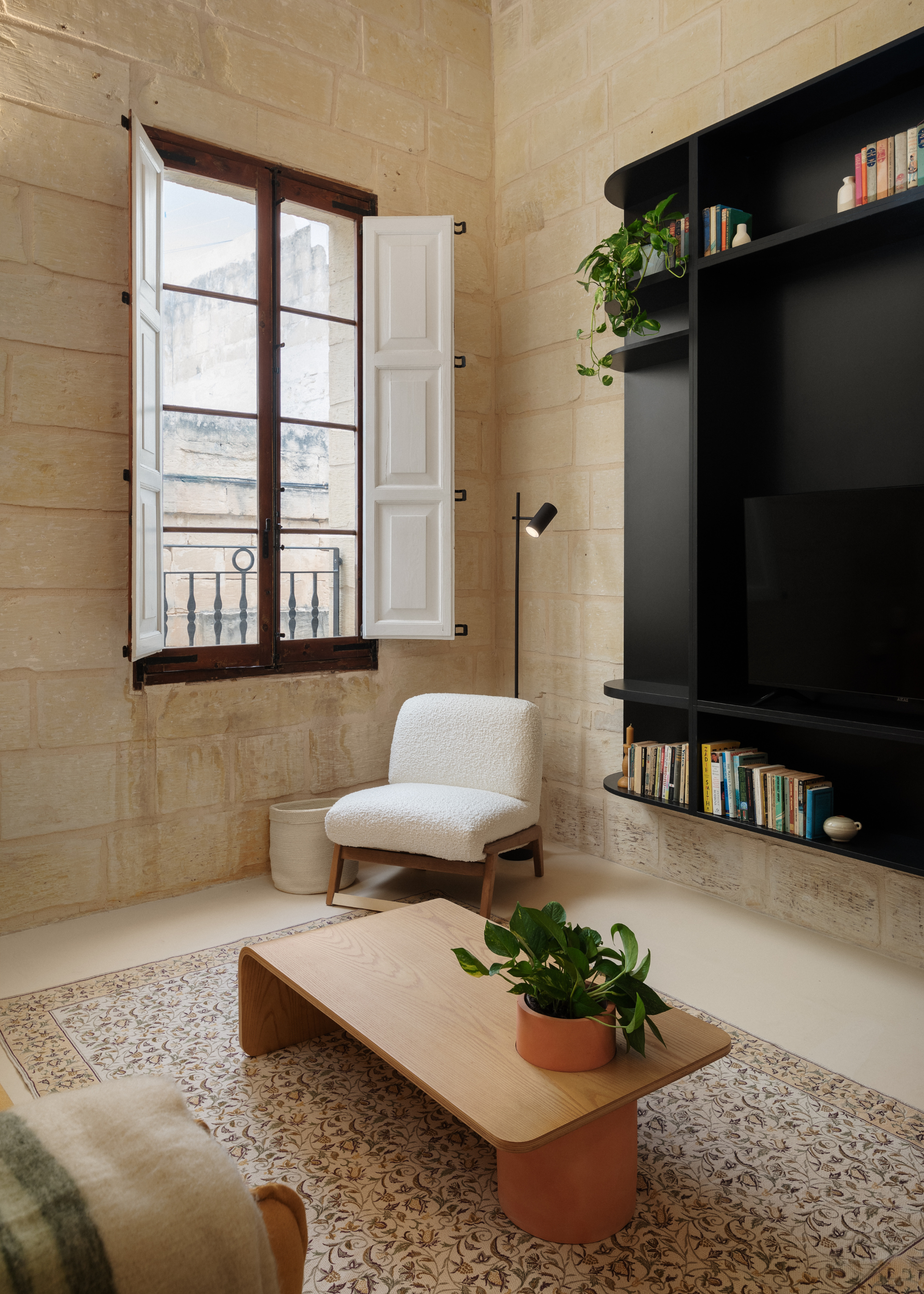 A living room with brick walls, a black wood unit for books, plants, and a TV, a tiled floor, a low wood table, a fuzzy wood sofa seater, and a black stem floor lamp