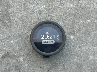 On a grey background a small black button size GPS sits with a battery indicator, a large digital clock and overall kilometers.