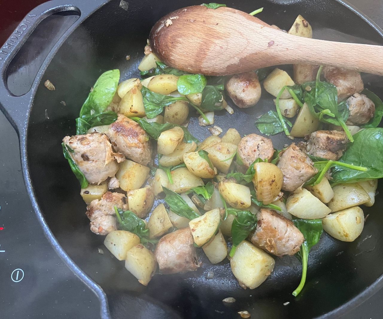 Chopped up sausages, potato, and spinach in a black cast iron pan with a wooden spoon