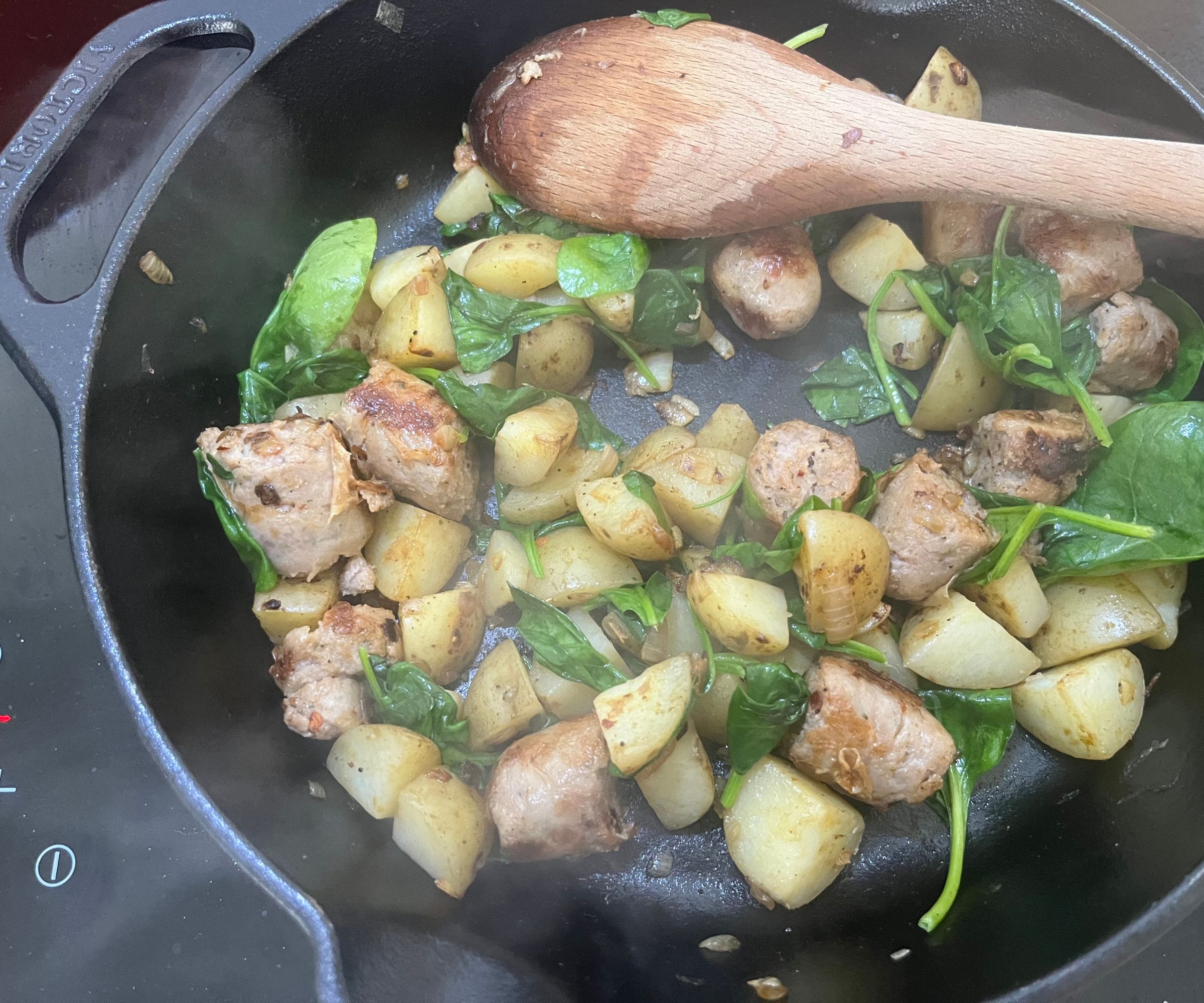 Chopped up sausages, potato, and spinach in a black cast iron pan with a wooden spoon