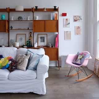 A living room with an off-white sofa and a rocking pink chair and a mid-century modern shelving unit in the background