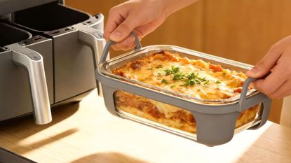 A glass air fryer dish with two silicone handles full of lasagne, being lifted away from an open Ninja air fryer