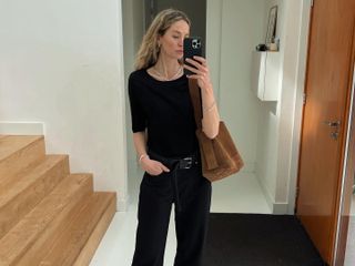 Fashion influencer Anouk Yve taking a mirror selfie wearing a chic fall outfit with belted jeans, black pointed heels, a short-sleeve black knit, and a suede bag.