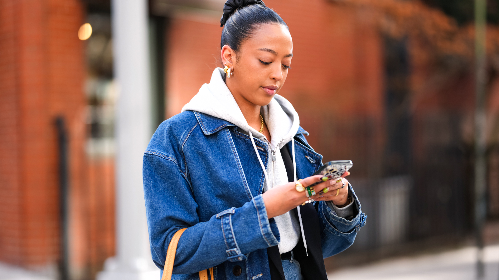 A woman stands in the street in front of brick buildings, the woman is wearing a grey hooded sweatshirt under a blue denim jacket and has several pretty rings on her fingers. The woman types a message on her phone.